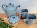 Jericho teapot, two mugs and a lidded honey pot by Matthew Freed on a wooden table, with a fern